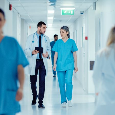 Surgeon and Female Doctor Walk Through Hospital Hallway, They Consult Digital Tablet Computer while Talking about Patient's Health. Modern Bright Hospital with Professional Staff.
